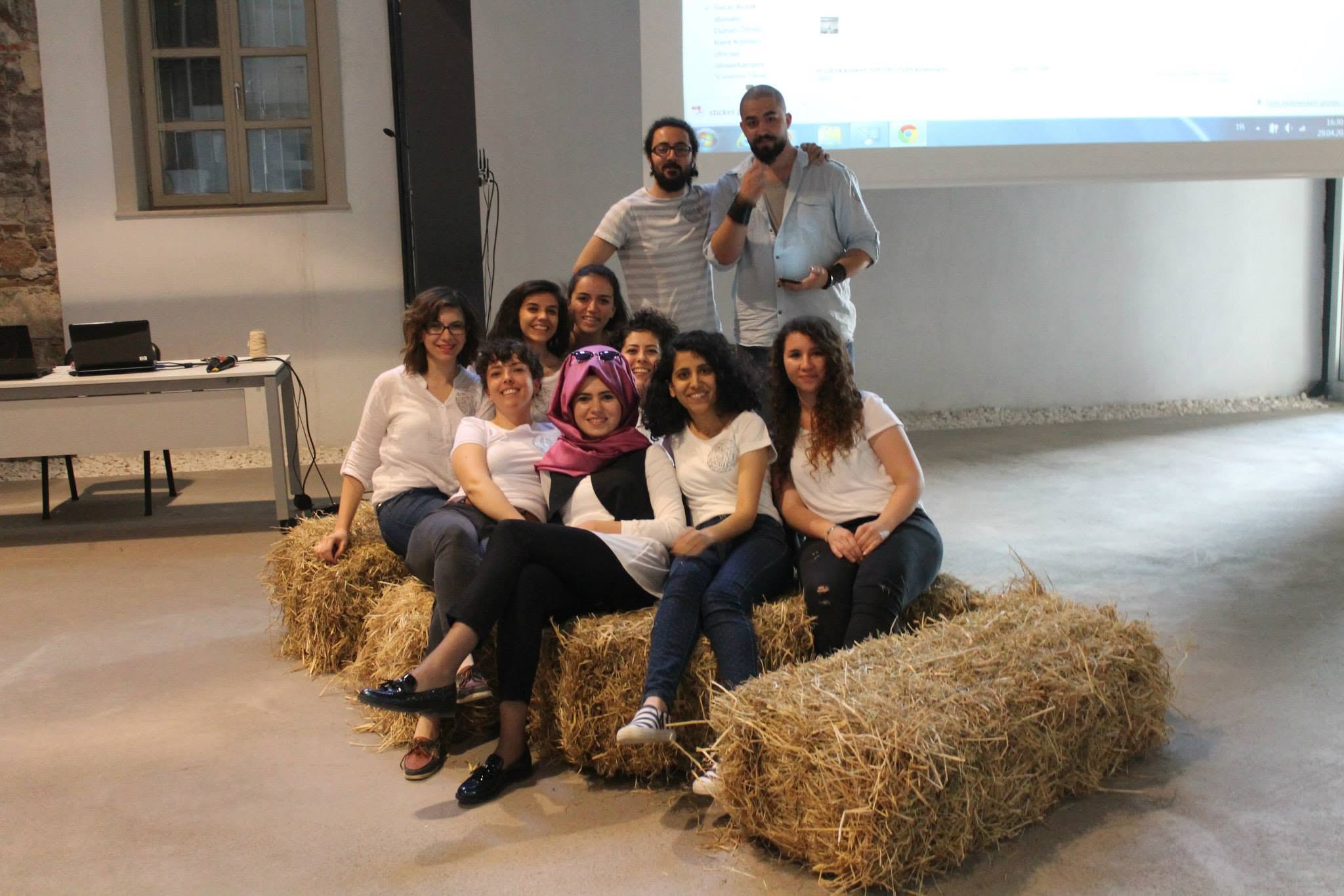 Organizing team meeting indoors, seated on hay bales in front of a presentation screen